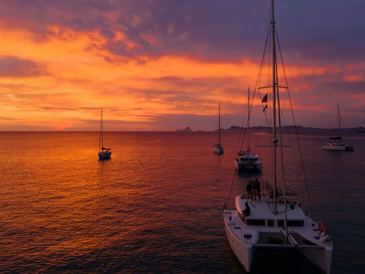 Catamarã e veleiros ancorados em mar calmo sob um pôr do sol alaranjado